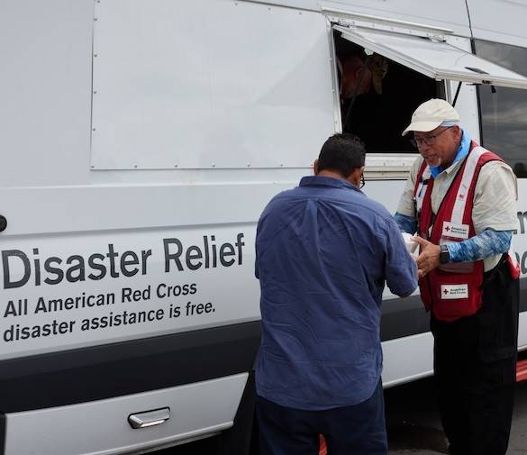 A Red Cross volunteer hands a client a warm meal in front of an emergency response vechicle showing the text "All American Red Cross dusaster assistance is free."