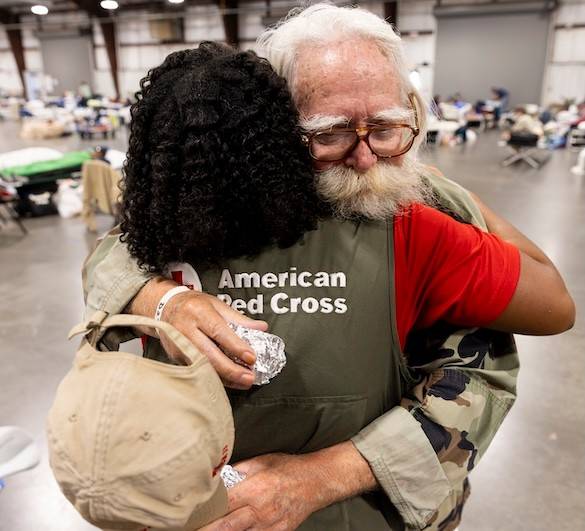 A dark haired Red Cross volunteer hugs an older gentleman with white hear and bushy white beard at a shelter.