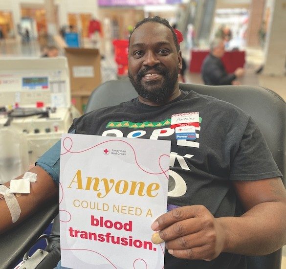 Smiling blood donor holds up a sign reading Anyone Could Need a Blood Transfusion